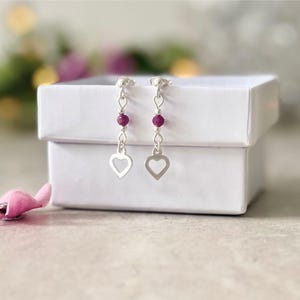 May include: A pair of silver drop earrings featuring a small, round, deep pink bead suspended above a silver heart-shaped charm. The earrings are displayed hanging from the edge of a plain white square gift box.