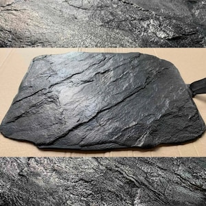 Puede incluir: Una bandeja de servir de pizarra negra con una superficie texturizada. La bandeja tiene un asa en el lateral.