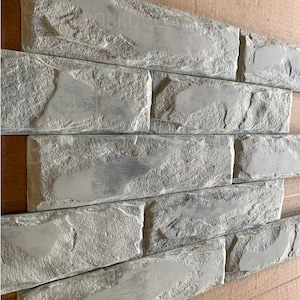 Puede incluir: Primer plano de una sección de paneles de ladrillo gris. El panel tiene una apariencia texturizada y desgastada, con formas de ladrillo rectangulares dispuestas en un patrón escalonado. El color es un gris apagado con reflejos más claros, lo que sugiere una textura similar a la piedra.