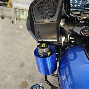 May include: A blue cylindrical cup holder mounted on a motorcycle fairing, containing a black and yellow Reign energy drink can. The motorcycle features a digital dashboard display, a black speaker grille, and a blue fuel tank.