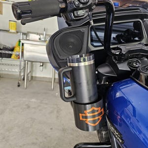 Può includere: Una tazza da viaggio nera con manico e coperchio trasparente, fissata in un portabicchieri nero con il logo arancione Harley-Davidson. Il portabicchieri è montato sul manubrio di una moto blu con comandi e specchietti di colore nero.