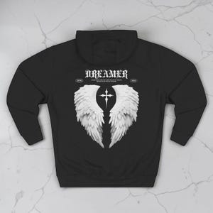 May include: Black hoodie with a white graphic of angel wings and a cross. The word "DREAMER" is printed above the wings in a gothic font. The hoodie has a hood and long sleeves.