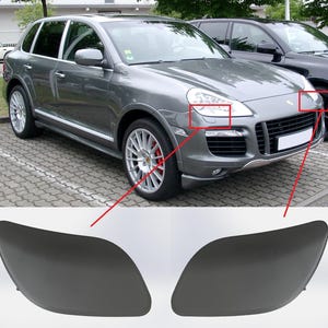 May include: A grey SUV with a black front bumper and black headlights. The car has a black grille and a black Porsche emblem. The car is parked on a grey asphalt surface.