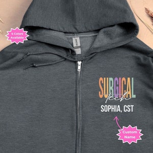 May include: Dark heather grey zip-up hoodie with a rainbow colored graphic that says "SURGICAL TECH" and "SOPHIA, CST" with a pink starburst that says "Custom Name" below.