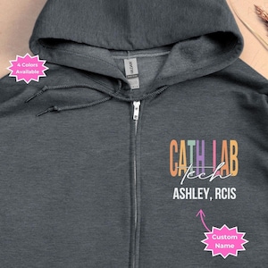 May include: Dark heather grey zip-up hoodie with a colorful "Cath Lab Tech" design and the text "Ashley, RCIS" below. The hoodie has a pocket on the front and a zipper closure.