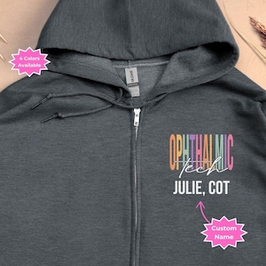 Op de afbeelding: Donkergrijze hoodie met rits en een regenboogkleurige afbeelding met de tekst "Ophthalmic Tech" en "Julie, COT". Onder de tekst staat een roze ster met de tekst "Custom Name".