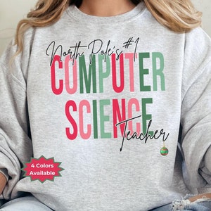 May include: A light gray sweatshirt with the text "North Pole's #1 COMPUTER SCIENCE Teacher" in red, green, and pink. A small Christmas ornament graphic is below the text. The words "4 Colors Available" are in a red and green starburst.