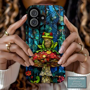 May include: A phone case featuring a stained-glass design with a frog sitting on a mushroom. The frog has red eyes and is green. The mushroom has a red, orange, and yellow top. The background is blue and green. The case is labeled "TOUGH CASE".