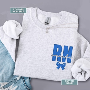 Könnte beinhalten: Ein hellgraues Sweatshirt mit den blauen Buchstaben "RN", dem Namen "amanda" in Schreibschrift und einer blauen Schleifen-Grafik. Der Text "5 FARBEN VERFÜGBAR" und "CUSTOM NAME" sind ebenfalls sichtbar. Eine silberne Halskette ist ebenfalls vorhanden.