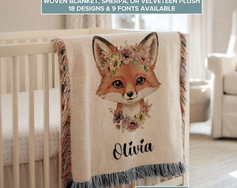 May include: A woven blanket with a fox design. The fox has a floral crown and is centered on the blanket. The name "Olivia" is printed below the fox. The blanket has a fringe border and is draped over a crib.