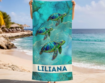 May include: A rectangular beach towel featuring a teal and blue marble background with three sea turtles in shades of green and blue. The name LILIANA is printed in blue capital letters on a white horizontal band near the bottom of the towel.