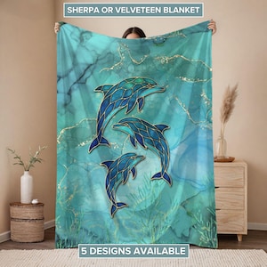 May include: A teal and blue blanket featuring three stylized dolphins with gold outlines. The blanket has a marble-like background with the text "SHERPA OR VELVETEEN BLANKET" and "5 DESIGNS AVAILABLE".
