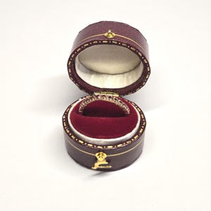 May include: A gold ring set with small red gemstones sits in an open, round, burgundy leather jewellery box. The box has a gold clasp and a red velvet interior. The ring is displayed against a red velvet background.