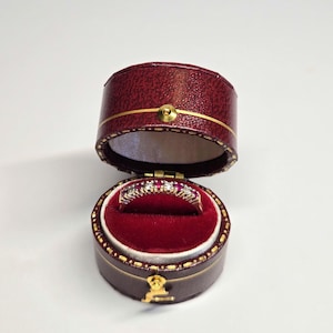 Peut inclure: Une bague en or sertie de pierres précieuses rouges et blanches, présentée dans un écrin en cuir bordeaux ouvert avec un fermoir doré. L'intérieur de la boîte est en velours rouge.
