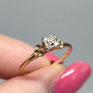 Könnte beinhalten: Ein Goldring mit einem quadratischen Mittelstück aus vier kleinen, klaren Steinen. Der Ring ist auf beiden Seiten der zentralen Fassung mit zwei kleinen, skulptierten Goldblumen verziert, was ein zartes und detailliertes Blumendesign ergibt.