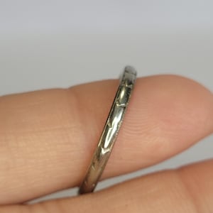 May include: A thin, metallic band ring featuring a repeating engraved pattern along its outer surface. The ring has a polished, reflective finish and a narrow profile, showcasing a series of small, uniform indentations that create a textured look.