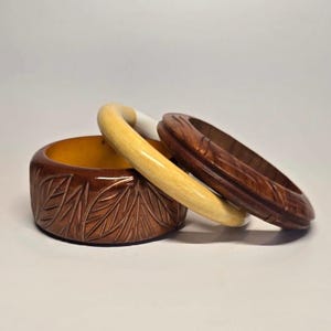 May include: A set of three stacked bangles including a wide dark brown wooden bracelet with a carved leaf pattern, a smooth light yellow wooden bangle, and a dark brown wooden bracelet featuring a central horizontal groove and carved lines.