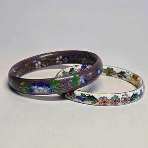 May include: Two round bangle bracelets featuring cloisonné enamel work. One bracelet has a dark purple base with blue, white, and green floral patterns. The second bracelet has a white base with multi-coloured floral designs and gold-tone metal edges.