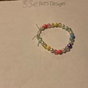 May include: A colorful beaded bracelet with white, pink, yellow, blue, and purple beads. The bracelet is made with small round beads and is strung on a clear elastic cord.