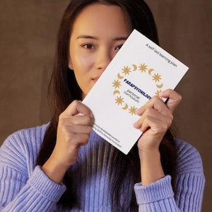 Puede incluir: Un folleto blanco titulado "PARAPSYCHOLOGY personal curriculum" con gráficos dorados de estrellas y lunas crecientes. El folleto se sostiene frente a una persona que lleva un suéter morado claro.