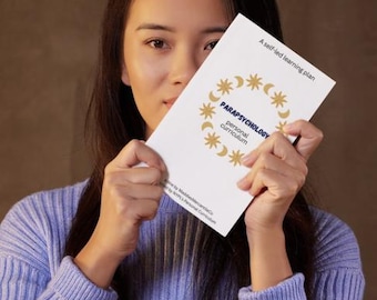 May include: A white booklet titled "PARAPSYCHOLOGY personal curriculum" with gold star and crescent moon graphics. The booklet is held in front of a person wearing a light purple sweater.