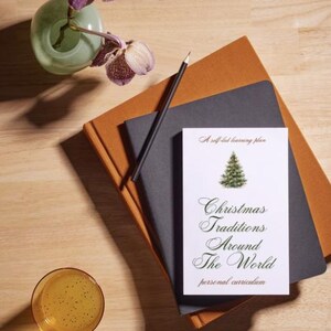 Puede incluir: Naturaleza muerta con una pila de cuadernos, un lápiz, un vaso de líquido y un jarrón con flores. El cuaderno superior tiene el texto "Christmas Traditions Around The World". La paleta de colores incluye marrón, negro y verde.