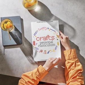 Puede incluir: Una persona sostiene un libro titulado "crafts personal curriculum" con un borde decorativo de suministros de artesanía. El libro está sobre una superficie gris junto a un cuaderno azul y un vaso de bebida con pajita. La persona lleva una camisa naranja.