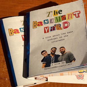 May include: A stack of zines titled "The Basement Yard" with colorful cut-out letters. The zine features a photo of three men and text that reads "a zine about the best podcast on the interwebs" and "a zine by MaddiesMercantileCo."