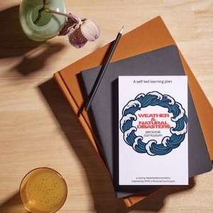 Puede incluir: Una pila de cuadernos con un lápiz y un libro titulado "Weather & Natural Disasters" con un diseño de olas. También hay un vaso de líquido amarillo y un jarrón con flores moradas.
