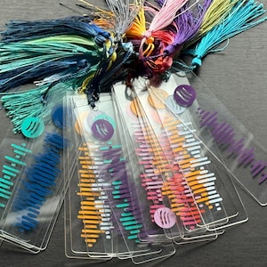 May include: A set of colourful acrylic bookmarks featuring a sound wave design and a small circle with a wave symbol. The bookmarks have tassels in various colours, including teal, purple, pink and orange.