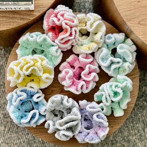 May include: A collection of colorful, handmade scrunchies in various pastel shades, including pink, yellow, blue, and green. Each scrunchie features a ruffled, textured design with a white trim, displayed on a wooden surface. These hair accessories are perfect for adding a touch of color to any hairstyle.