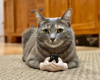 Könnte beinhalten: Eine graue Tabby-Katze mit weißen Pfoten liegt auf einem Teppich. Die Katze hält ein schwarzes Smartphone in ihren beiden weißen, menschenähnlichen Händen.
