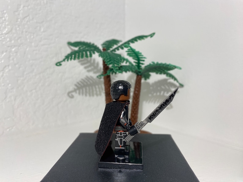 Op de afbeelding: Een zwart Lego minifiguur met een zwarte cape en een zilveren zwaard. Het minifiguur staat op een zwarte basis.