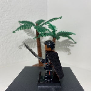 Op de afbeelding: Een zwart Lego minifiguur met een zwarte cape en een zilveren zwaard. Het minifiguur heeft een zwart haarstukje en een bruin gezicht. Het minifiguur staat op een zwarte basis.