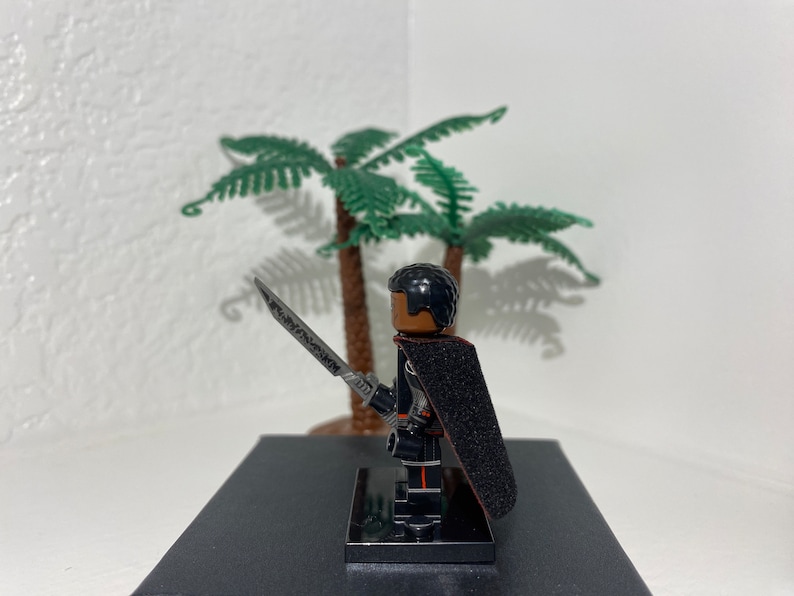 Op de afbeelding: Een zwart Lego minifiguur met een zwarte cape en een zilveren zwaard. Het minifiguur heeft een zwart haarstukje en een bruin gezicht. Het minifiguur staat op een zwarte basis.
