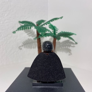 Op de afbeelding: Een zwart Lego minifiguur met een zwarte cape en een zwart hoofd met een textuur. Het minifiguur staat op een zwarte basis voor een groene palmboom.