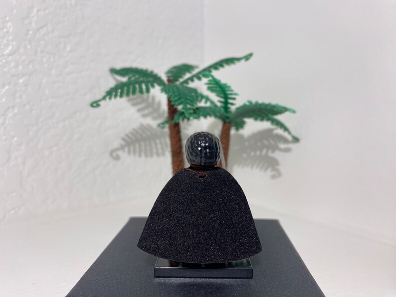 Op de afbeelding: Een zwart Lego minifiguur met een zwarte cape en een zwart hoofd met een textuur. Het minifiguur staat op een zwarte basis voor een groene palmboom.