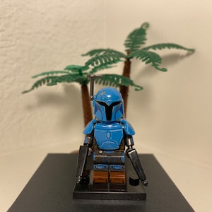 May include: A blue and black Lego minifigure of a Mandalorian warrior with a blaster rifle in each hand. The minifigure is standing on a black base.