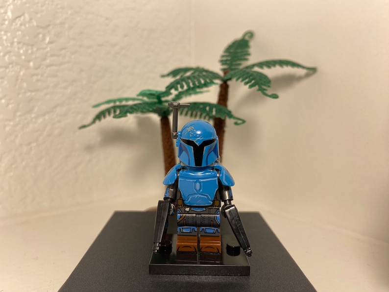May include: A blue and black Lego minifigure of a Mandalorian warrior with a blaster rifle in each hand. The minifigure is standing on a black base.