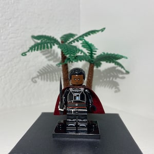 Op de afbeelding: Een zwart Lego minifiguur met een rode cape en een zwaard. Het minifiguur heeft een donkerbruine huidskleur en een serieuze uitdrukking. Het minifiguur staat op een zwarte basis.
