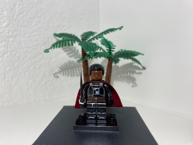 Op de afbeelding: Een zwart Lego minifiguur met een rode cape en een zwaard. Het minifiguur heeft een donkerbruine huidskleur en een serieuze uitdrukking. Het minifiguur staat op een zwarte basis.