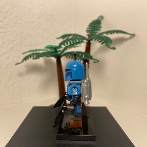 May include: A blue and gray Lego minifigure of a Mandalorian warrior with a jetpack and a blaster rifle. The minifigure is standing on a black base.