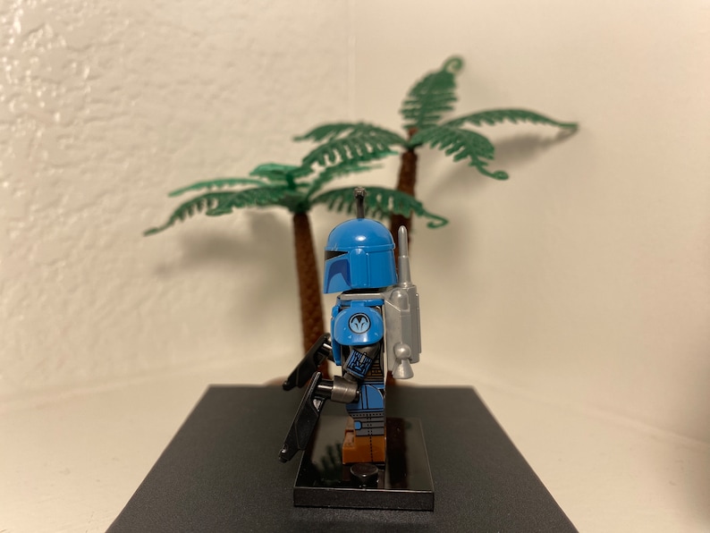 May include: A blue and gray Lego minifigure of a Mandalorian warrior with a jetpack and a blaster rifle. The minifigure is standing on a black base.