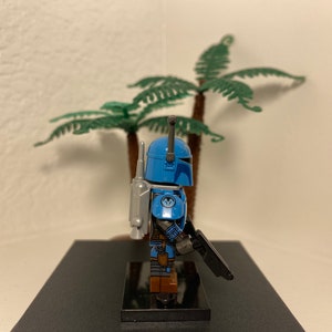 May include: A blue and gray Lego minifigure of a Mandalorian with a jetpack and a blaster rifle. The minifigure is standing on a black base.