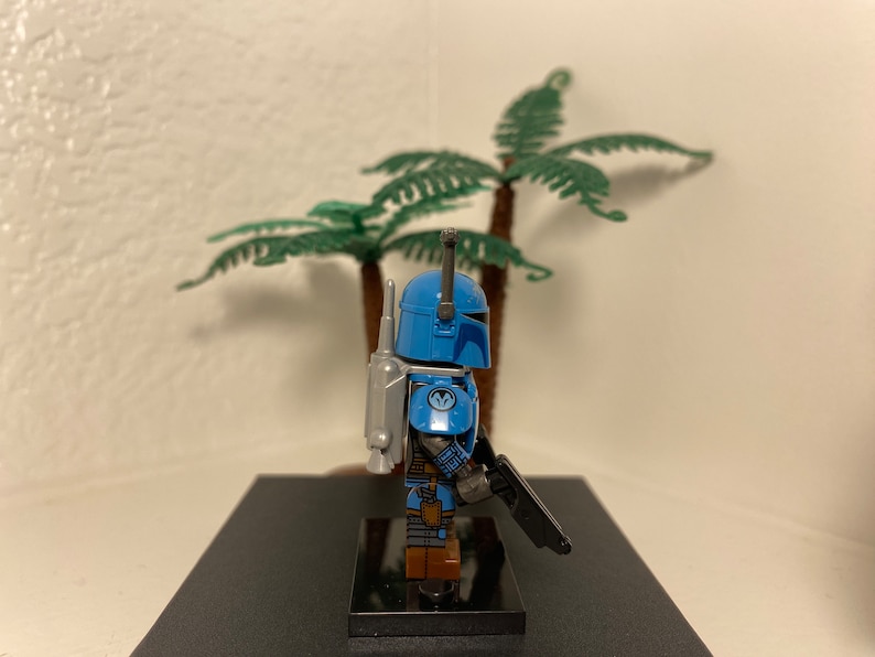 May include: A blue and gray Lego minifigure of a Mandalorian with a jetpack and a blaster rifle. The minifigure is standing on a black base.
