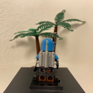 May include: A blue and gray Lego minifigure of a Star Wars character, Boba Fett, standing on a black base. The minifigure is wearing a blue helmet and armor with a gray jetpack.