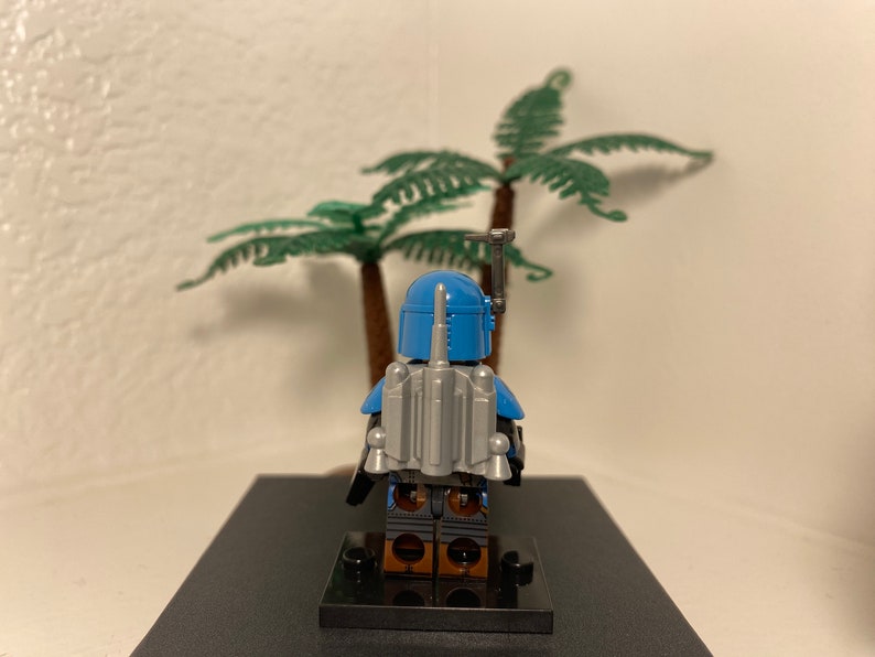 May include: A blue and gray Lego minifigure of a Star Wars character, Boba Fett, standing on a black base. The minifigure is wearing a blue helmet and armor with a gray jetpack.