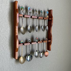 May include: A wooden spoon rack with two rows of decorative spoons. The spoons have various designs on the handles and are made of different metals. The rack is made of dark brown wood and is mounted on a light-colored wall.