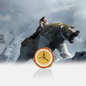 May include: A young girl rides a large, white, armoured polar bear through a snowy mountain landscape. The bear is wearing a golden helmet and armour. The girl is wearing a brown coat and a white scarf. A compass with a gold and white design is in the foreground.
