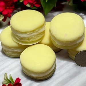 May include: Five yellow macarons with white filling. The macarons are arranged in a pile on a white surface.